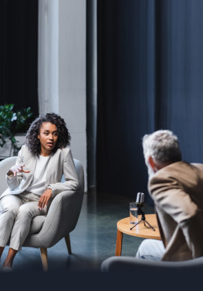 african american journalist gesturing while talking with blurred businessman sitting in armchair during interview