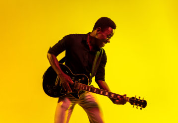 Young african-american musician playing the guitar like a rockstar on yellow background